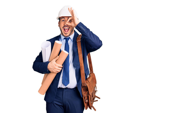 Jeune homme hispanique portant un casque d'architecte et un sac en cuir tenant des plans souriants heureux faisant signe ok avec la main sur les yeux en regardant à travers les doigts  - Photo, image