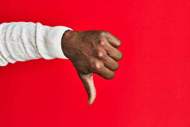 Χέρι και χέρι του αφροαμερικανού μαύρου άνδρα πάνω από το κόκκινο απομονωμένο φόντο κάνει αντίχειρες προς τα κάτω χειρονομία απόρριψης, αντιπάθεια αποδοκιμασίας και αρνητικό σημάδι  - Φωτογραφία, εικόνα