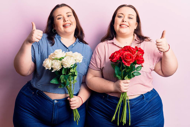 Young plus size twins holding flowers smiling happy and positive, thumb up doing excellent and approval sign  - Zdjęcie, obraz