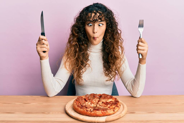 jong hispanic meisje eten smakelijke pepperoni pizza maken vis gezicht met mond en squinting ogen, gek en komisch.  - Foto, afbeelding