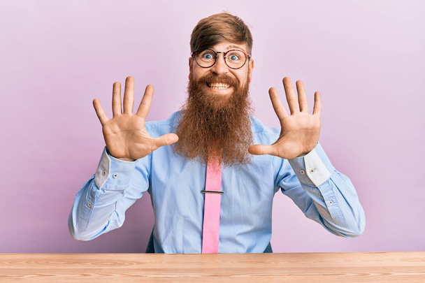 Молодой ирландский рыжий мужчина в деловой рубашке и галстуке сидит на столе и показывает пальцами номер десять, улыбаясь уверенно и счастливо.  - Фото, изображение