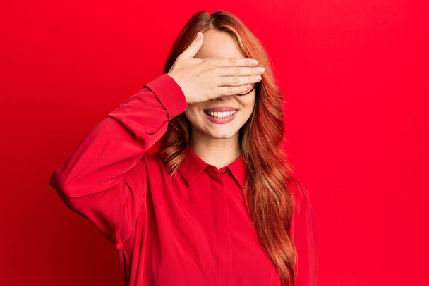 Fiatal gyönyörű vörös hajú nő visel alkalmi ruhák és szemüveg felett piros háttér mosolyog és nevet a kezét az arcon takaró szemek meglepetés. vak fogalom.  - Fotó, kép