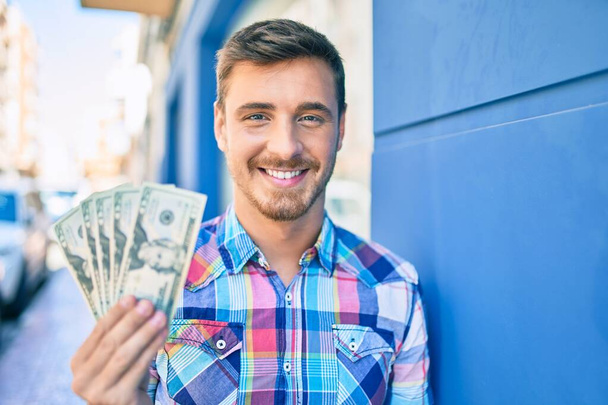 Young caucasian man smiling happy holding dollars banknotes leaning on the wall at the city. - Foto, afbeelding