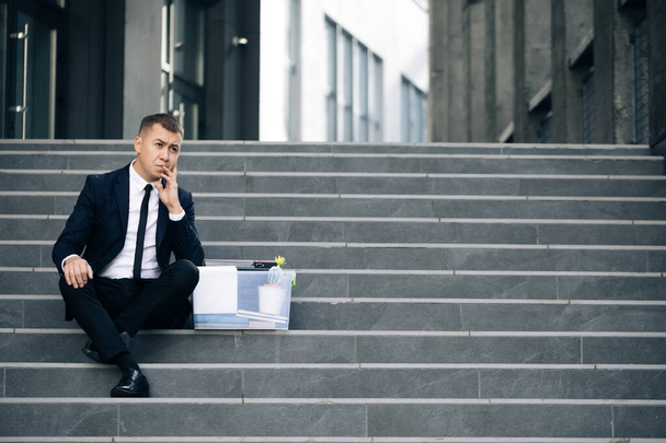 Sad businessman sitting on stairs outdoor with box of stuff as lost business. Fired man. Unemployment rate growing due pandemic. Male office worker in despair lost job - Fotografie, Obrázek