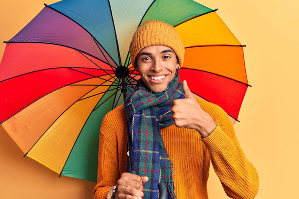 Young african amercian man holding colorful umbrella smiling happy and positive, thumb up doing excellent and approval sign  - Photo, Image