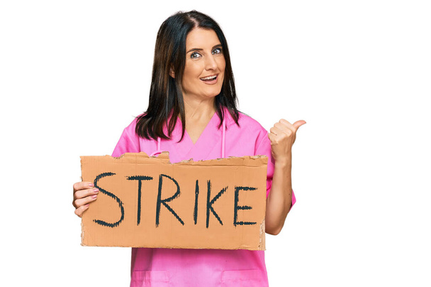 Брюнетка-лікар середнього віку жінка тримає страйк банер картон вказуючи великий палець в сторону посміхаючись щасливий з відкритим ротом
  - Фото, зображення