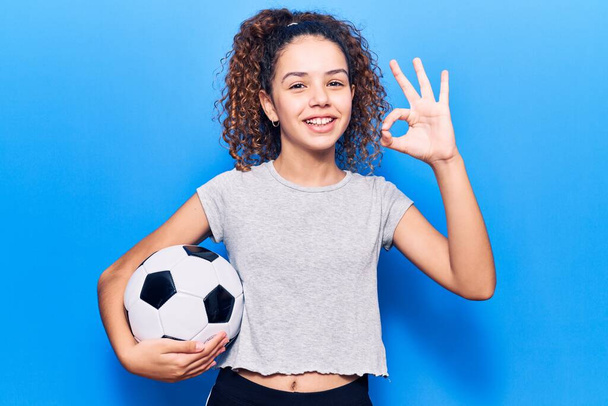 Krásná dívka s kudrnaté vlasy drží fotbalový míč dělá v pořádku znamení s prsty, usmívá přátelské gestikulace vynikající symbol  - Fotografie, Obrázek