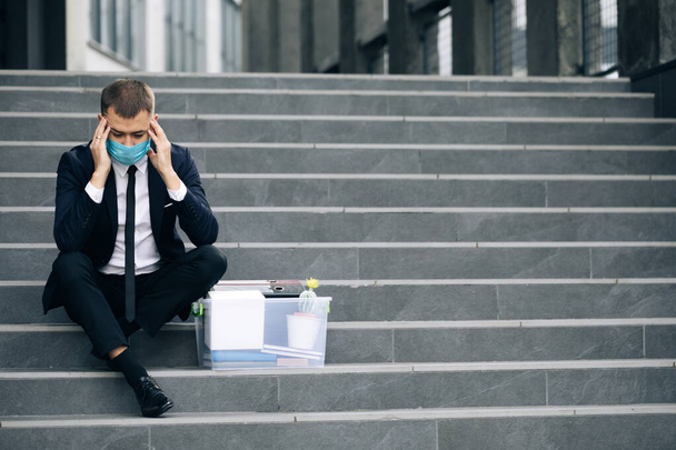 Fired male office worker in medical mask sitting on stairs in depression with box of stuff. Anxious concept. Workless man in despair. Unemployed businessman lost his business - Zdjęcie, obraz