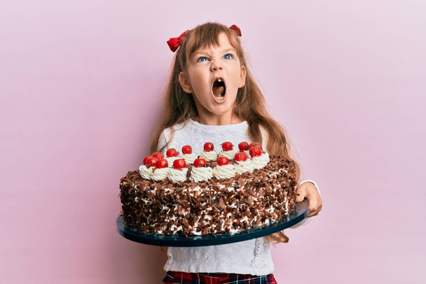 Маленькая кавказская девочка празднует день рождения, держа в руках большой шоколадный торт злой и безумный кричащий расстроенный и разъяренный, кричащий от гнева. гнев и агрессивная концепция.  - Фото, изображение
