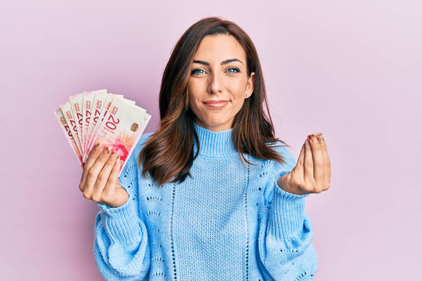 Młoda brunetka dzierżąca 20 sztabek banknotów izraelskich wykonujących gesty pieniężne dłońmi, prosząca o wypłatę pensji, biznes milionera  - Zdjęcie, obraz