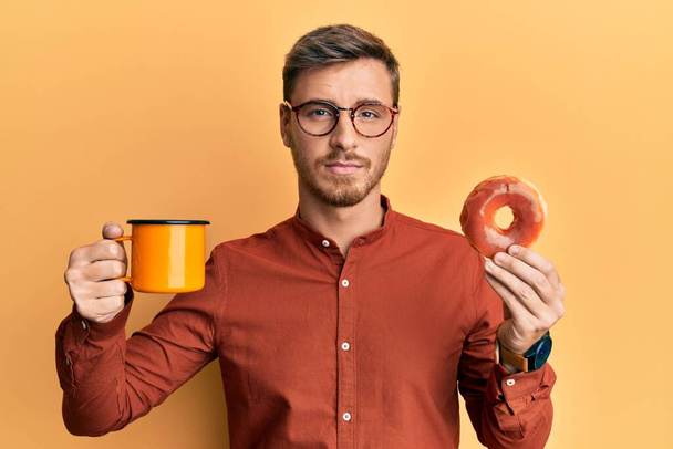 Knappe blanke man die donut eet en koffie drinkt ontspannen met een serieuze uitdrukking op zijn gezicht. eenvoudig en natuurlijk kijkend naar de camera.  - Foto, afbeelding