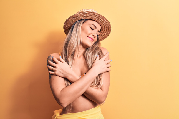 Joven mujer rubia hermosa con bikini y sombrero de verano sobre fondo amarillo aislado abrazándose feliz y positivo, sonriendo confiado. Amor propio y cuidado personal - Foto, imagen