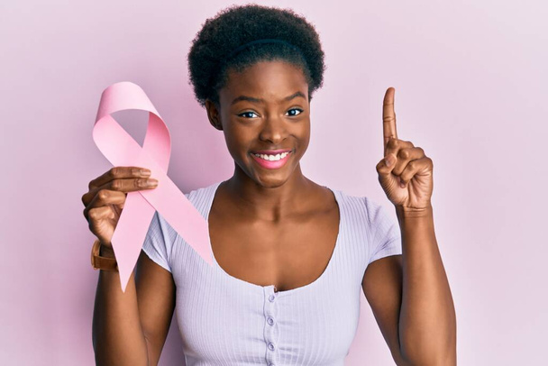 Young african american girl holding pink cancer ribbon surprised with an idea or question pointing finger with happy face, number one  - Fotografie, Obrázek