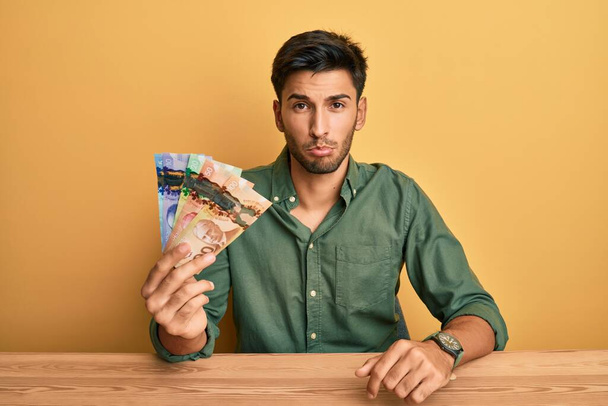 Mladý pohledný muž držící kanadské dolary v depresi a strachující se o úzkost, rozzlobený a vystrašený. smutný výraz.  - Fotografie, Obrázek