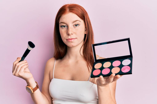 Young red head girl holding makeup brush and blush palette relaxed with serious expression on face. simple and natural looking at the camera.  - Photo, Image