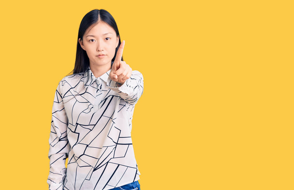 Młoda piękna Chinka w luźnej koszuli wskazującej palcem w górę i gniewnym wyrazie twarzy, nie pokazująca gestu  - Zdjęcie, obraz
