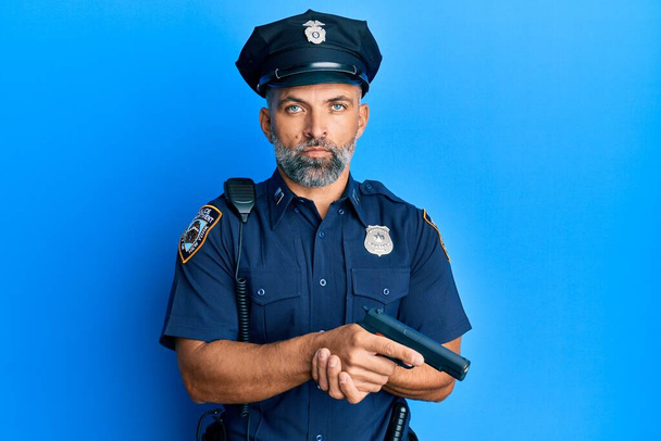 Hombre guapo de mediana edad con uniforme de policía sosteniendo pistola relajado con expresión seria en la cara. simple y natural mirando a la cámara.  - Foto, imagen