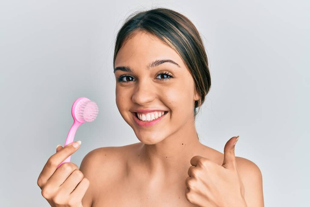 jonge brunette vrouw met behulp van gezicht scrubben borstel glimlachen gelukkig en positief, duim omhoog doen uitstekend en goedkeuring teken  - Foto, afbeelding