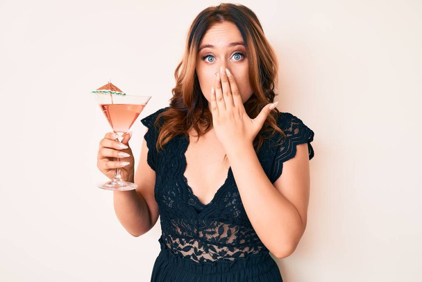 Νεαρή όμορφη Καυκάσια γυναίκα πίνει τροπικό κοκτέιλ με αλκοόλ που καλύπτει το στόμα με το χέρι, σοκαρισμένη και φοβισμένη για λάθος. έκπληκτη έκφραση  - Φωτογραφία, εικόνα