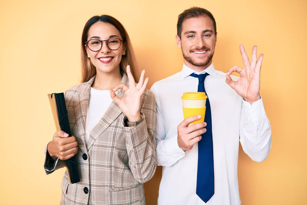 Beautiful couple wearing business clothes drinking take away coffee holding folder doing ok sign with fingers, smiling friendly gesturing excellent symbol  - Fotoğraf, Görsel