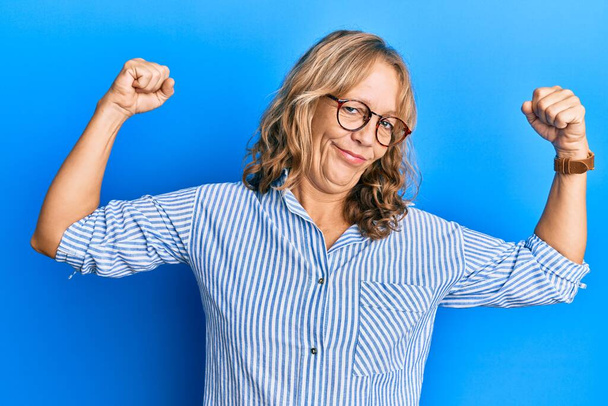 Mujer rubia de mediana edad que usa ropa casual y gafas que muestran los músculos de los brazos sonriendo orgullosos. concepto de fitness.  - Foto, Imagen