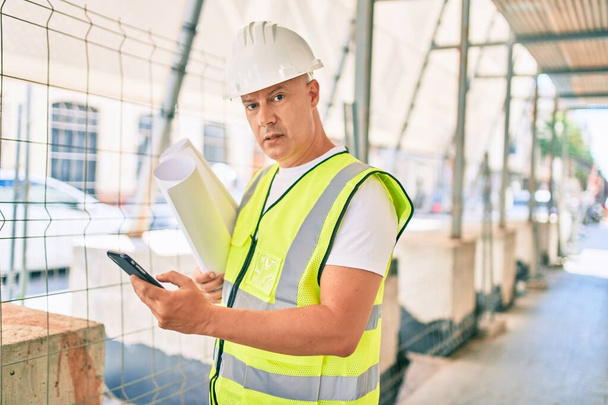 Middle age architect man with serious expression using smartphone and holding blueprints at the city. - Φωτογραφία, εικόνα