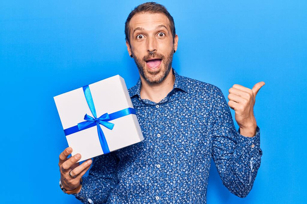 Молодой красивый мужчина держит в руках подарок указывая пальцем вверх в сторону улыбаясь счастливо с открытым ртом  - Фото, изображение