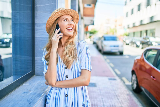 Yaz şapkası takmış, mutlu bir şekilde gülümseyen güzel sarışın bir kadın. Yüzünde gülümsemeyle duvara yaslanarak şehrin caddesindeki akıllı telefondan konuşarak.. - Fotoğraf, Görsel