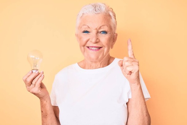 Старшая красивая женщина с голубыми глазами и седыми волосами держит лампочку для вдохновения и идеи улыбаясь с идеей или вопросом указывая пальцем со счастливым лицом, номер один  - Фото, изображение