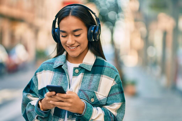 Молода латинська дівчина посміхається щаслива, використовуючи смартфон і навушники в місті
. - Фото, зображення