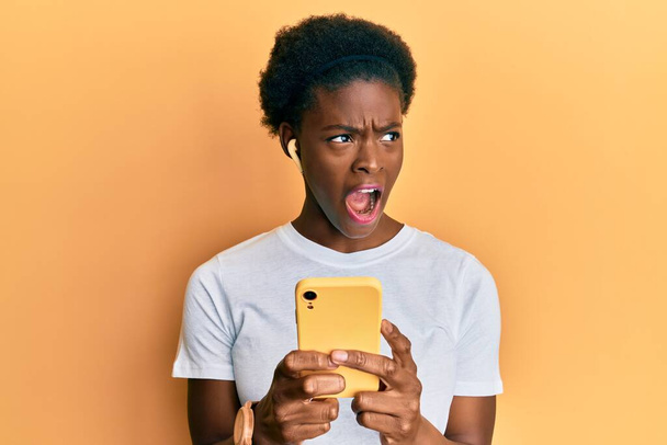Jovem afro-americana usando smartphones e fones de ouvido com raiva e gritos loucos frustrados e furiosos, gritando com raiva. raiva e conceito agressivo.  - Foto, Imagem
