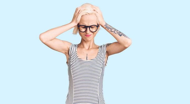 Молодая блондинка с татуировкой в повседневной одежде и очках страдает от головной боли в отчаянии и стрессе из-за боли и мигрени. руки на голове.  - Фото, изображение