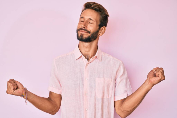 Handsome caucasian man with beard wearing casual pink shirt very happy and excited doing winner gesture with arms raised, smiling and screaming for success. celebration concept.  - Photo, Image