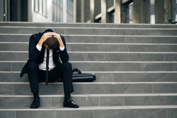 Tired putting hands to the head. Male office worker in despair lost job. Feeling bad and annoyed. Sad young businessman sitting on the stairs near office building - 写真・画像