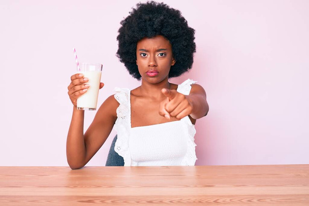 Giovane donna afro-americana che tiene un bicchiere di latte puntato con il dito verso la macchina fotografica e verso di te, gesto fiducioso che sembra serio  - Foto, immagini