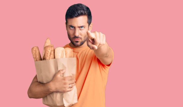 Young hispanic man holding paper bag with bread pointing with finger to the camera and to you, confident gesture looking serious  - Valokuva, kuva