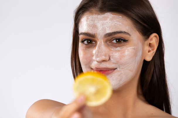 Enlarged photo. The brunette with facial mask smiles and holds out a slice of lemon to the camera. High quality photo - Foto, Imagen