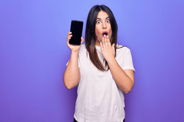 Giovane bella donna bruna che tiene smartphone mostrando schermo su sfondo viola che copre la bocca con la mano, scioccata e spaventata per errore. Espressione sorpresa - Foto, immagini