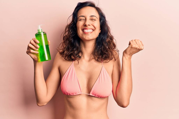 Young beautiful hispanic woman wearing bikini holding aloe vera lotion screaming proud, celebrating victory and success very excited with raised arm  - 写真・画像