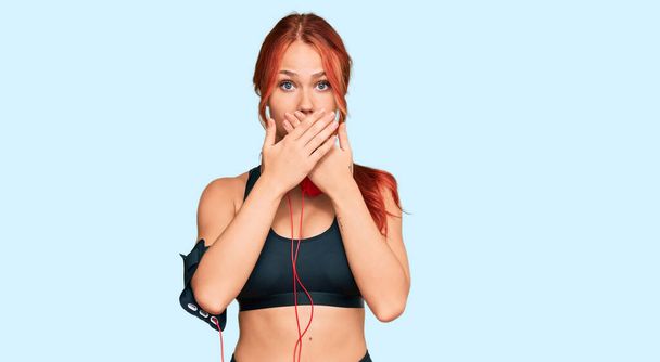 Young redhead woman wearing gym clothes and using headphones shocked covering mouth with hands for mistake. secret concept.  - Photo, Image