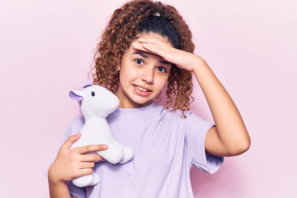 Hermosa niña con pelo rizado sosteniendo juguete muñeca animal estresado y frustrado con la mano en la cabeza, cara sorprendida y enojada  - Foto, Imagen