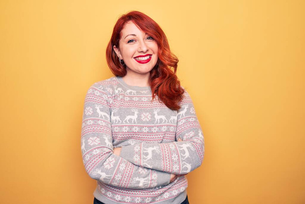 Schöne rothaarige Frau im Weihnachtspullover mit Rentieren über gelbem Hintergrund, fröhliches Gesicht, lächelnd mit verschränkten Armen, die in die Kamera schauen. Positiver Mensch. - Foto, Bild