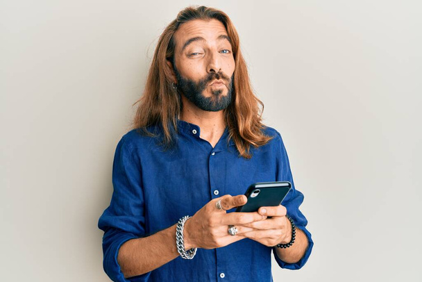 Attractive man with long hair and beard using smartphone looking at the camera blowing a kiss being lovely and sexy. love expression.  - Foto, Imagem