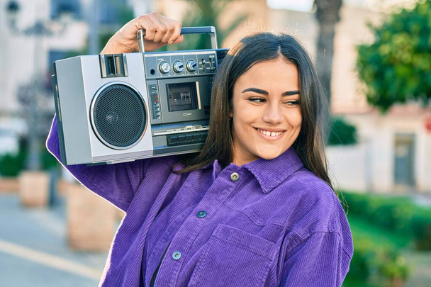 Jonge Spaanse vrouw glimlachend gelukkig luisterend naar muziek met boombox in de stad. - Foto, afbeelding