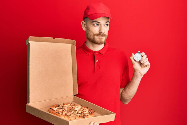 Молодой рыжий мужчина в униформе курьера держит коробку пиццы и часы обратного отсчета улыбаются, глядя в сторону и думая:.  - Фото, изображение