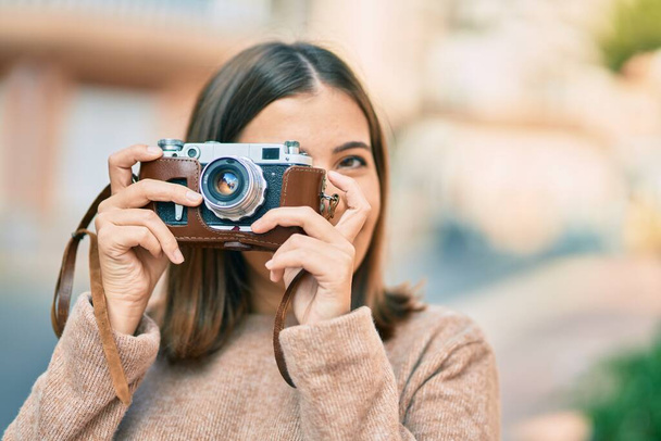 Νεαρή Ισπανίδα τουρίστρια χαμογελά ευτυχισμένη χρησιμοποιώντας παλιά κάμερα στην πόλη. - Φωτογραφία, εικόνα