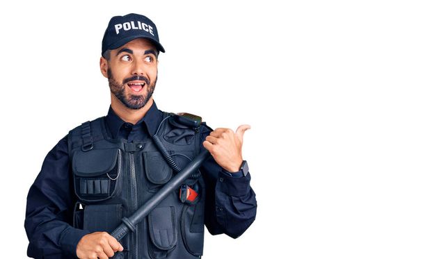 Young hispanic man wearing police uniform holding baton pointing thumb up to the side smiling happy with open mouth  - Photo, Image