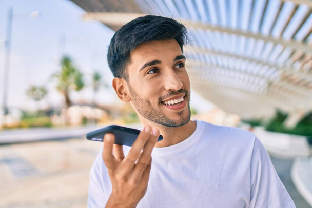 Giovane uomo latino sorridente felice invio di messaggi vocali utilizzando smartphone in città. - Foto, immagini