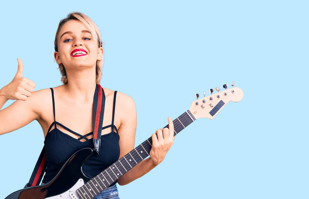 Genç ve güzel sarışın kadın elektronik gitar çalıyor mutlu ve pozitif gülümsüyor, başparmağıyla mükemmel ve onay işareti yapıyor.  - Fotoğraf, Görsel