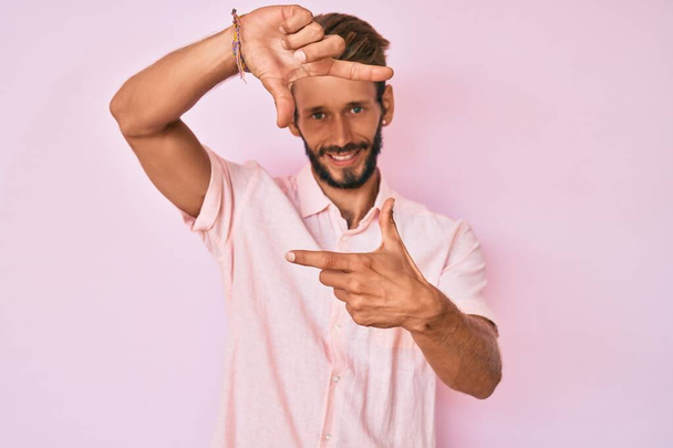 ピンクのカジュアルなシャツを着て髭を生やしたハンサムな白人男性は、幸せな顔をした手と指でフレームを作って笑っています。創造性と写真の概念.  - 写真・画像